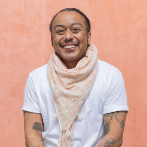 Mason, a nonbinary trans man with medium-brown skin, a nosering, and a moustache sits in front of a peach colored wall. they wear a plain white tshirt and peach-and-white keffiye. they have tattoos on their arms.